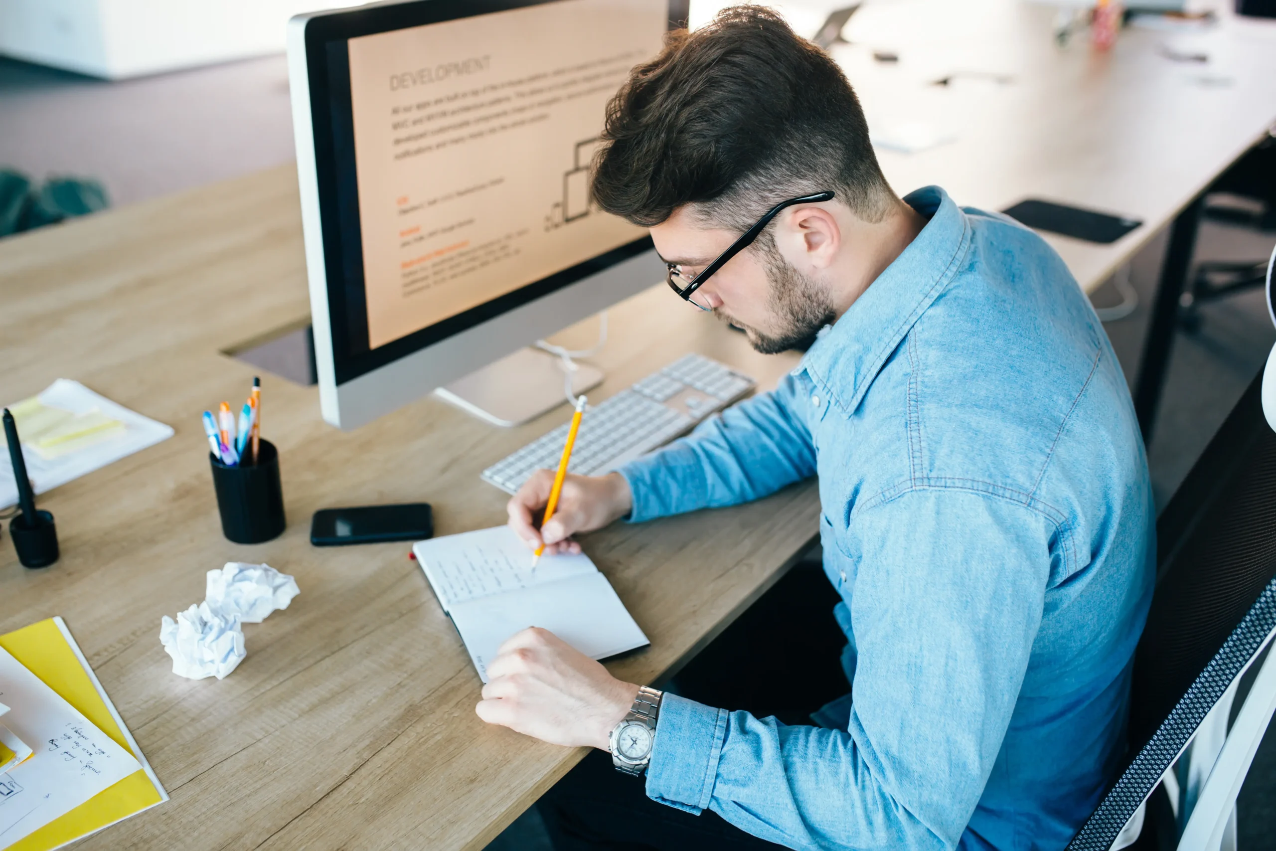 young-man-glassess-is-working-his-workplace-office-he-wears-blue-shirt-he-is-writing-notebook-view-from